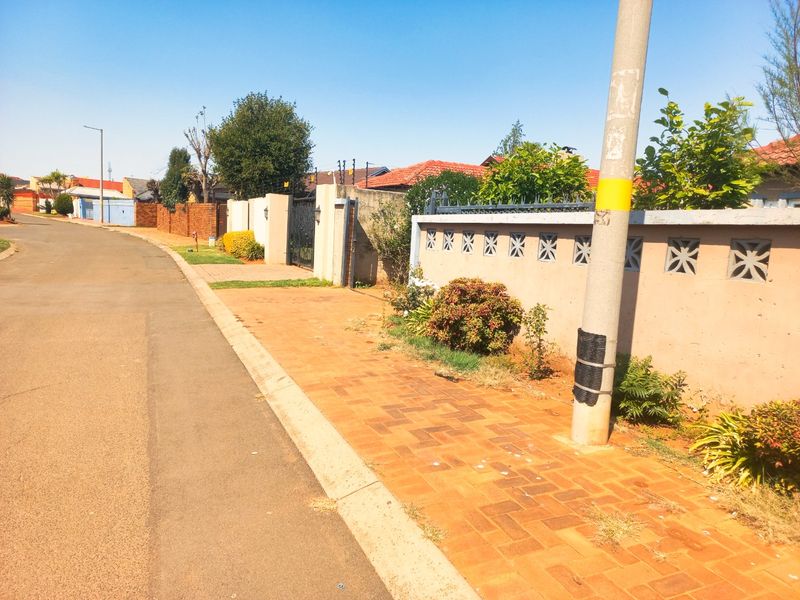 A street-level view of a residential property featuring a paved sidewalk, perimeter boundary wall with decorative blocks, and electric fencing.