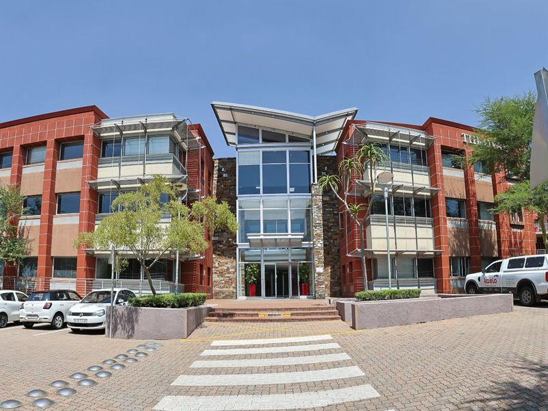 A modern commercial office building featuring a central glass atrium, stone facade accents, and an extensive paved parking area with pedestrian access.