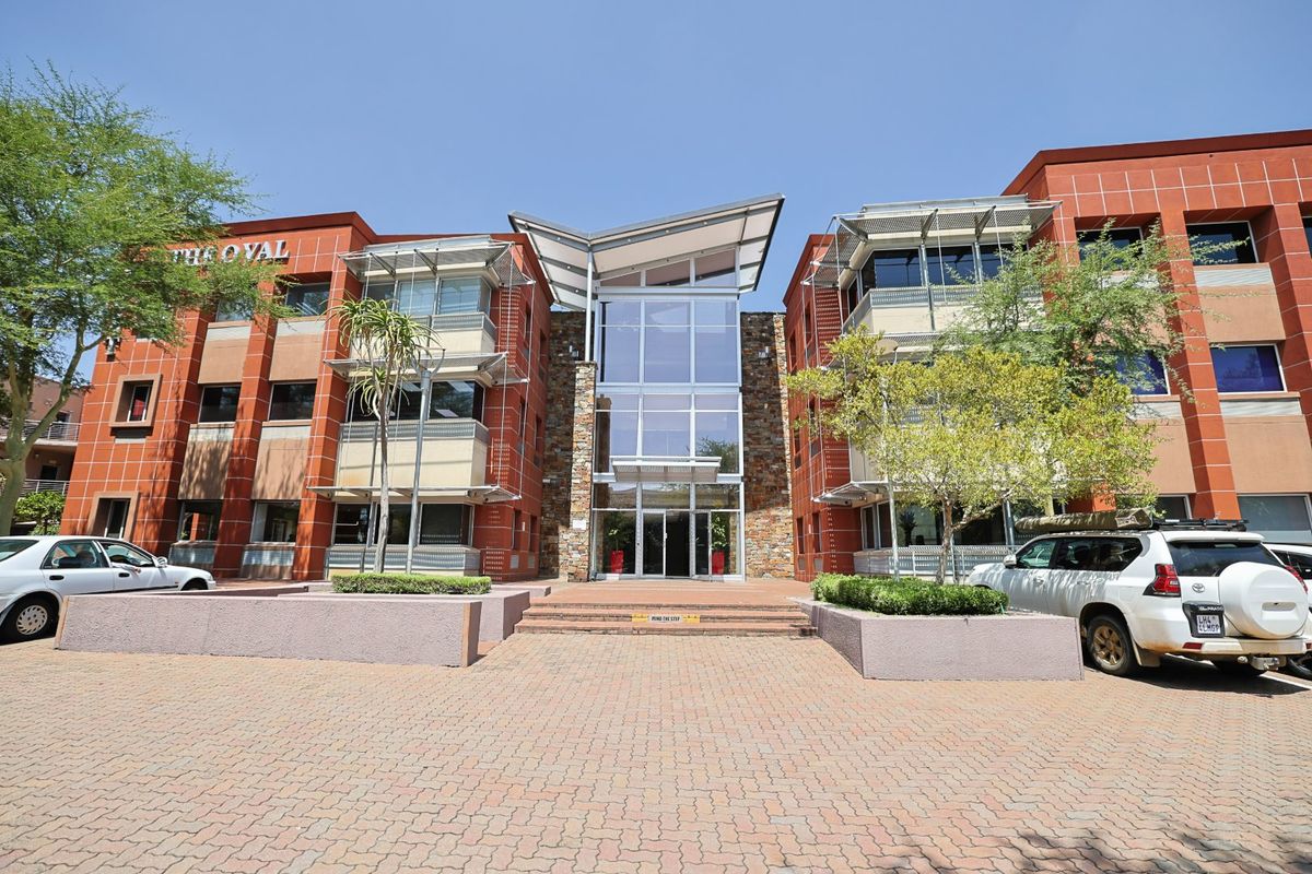 Front elevation of a modern commercial office building featuring a prominent glass atrium entrance, stone facade accents, and paved forecourt.
