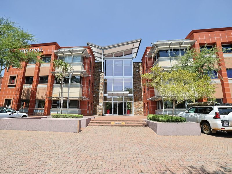 Front elevation of a modern commercial office building featuring a prominent glass atrium entrance, stone facade accents, and paved forecourt.