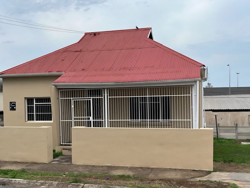 Front elevation of a single-story house featuring a red corrugated metal roof, security bars on windows, and a low perimeter boundary wall.