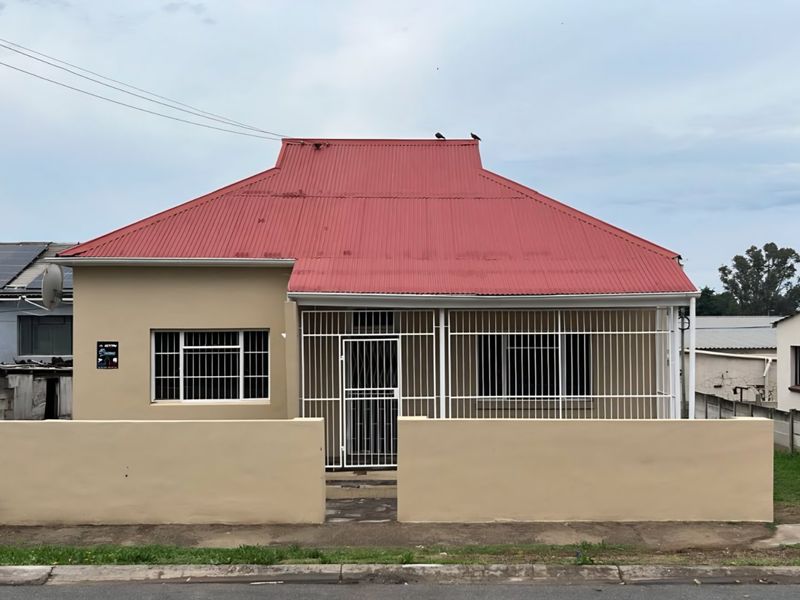 Front elevation of a single-story house featuring a red corrugated metal roof, security bars on windows, and a solid perimeter wall with a security gate.