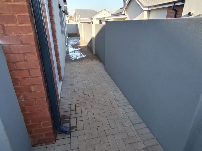 A narrow paved side passage between the house and the boundary wall, featuring a brick facade and drainage downpipe.