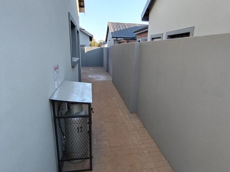 A clean, paved side passage between two residential buildings featuring a secure gas cylinder storage cage and modern boundary walling.