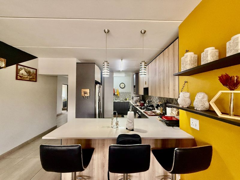 Modern kitchen featuring a central island with breakfast bar, wood-finish cabinetry, integrated appliances, and a vibrant yellow accent wall with floating shelving.