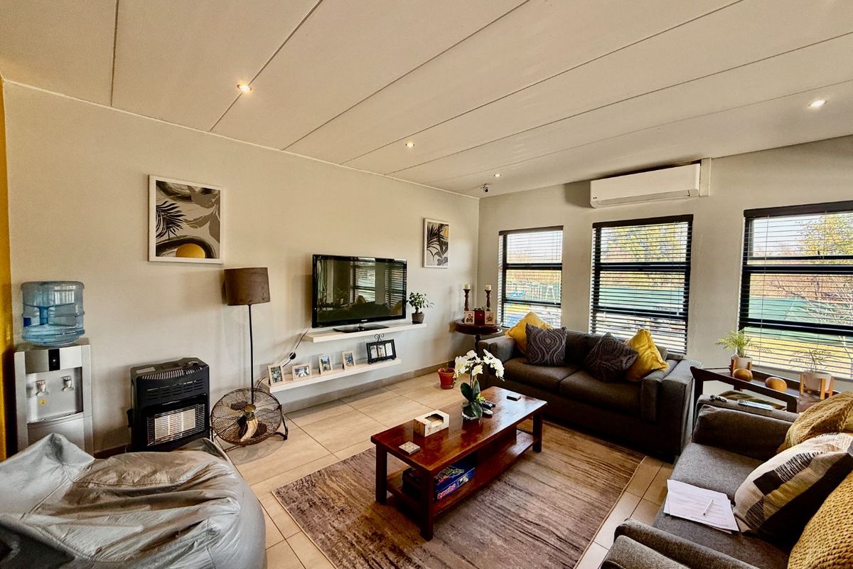 A well-lit living room featuring tiled flooring, recessed ceiling lights, a wall-mounted air conditioning unit, and large windows with venetian blinds.