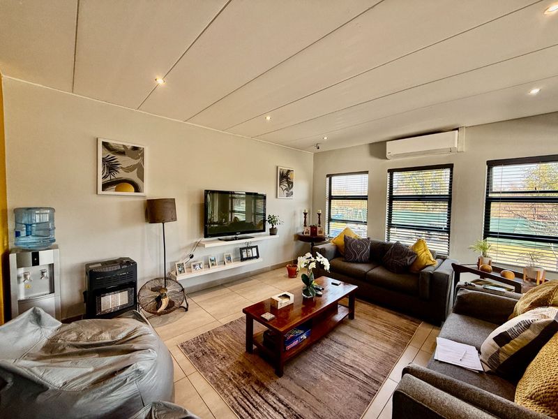 A well-lit living room featuring tiled flooring, recessed ceiling lights, a wall-mounted air conditioning unit, and large windows with venetian blinds.