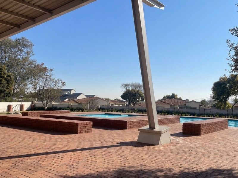 A well-maintained communal pool area featuring brick paving, built-in brick seating, and a covered patio structure under a clear sky.