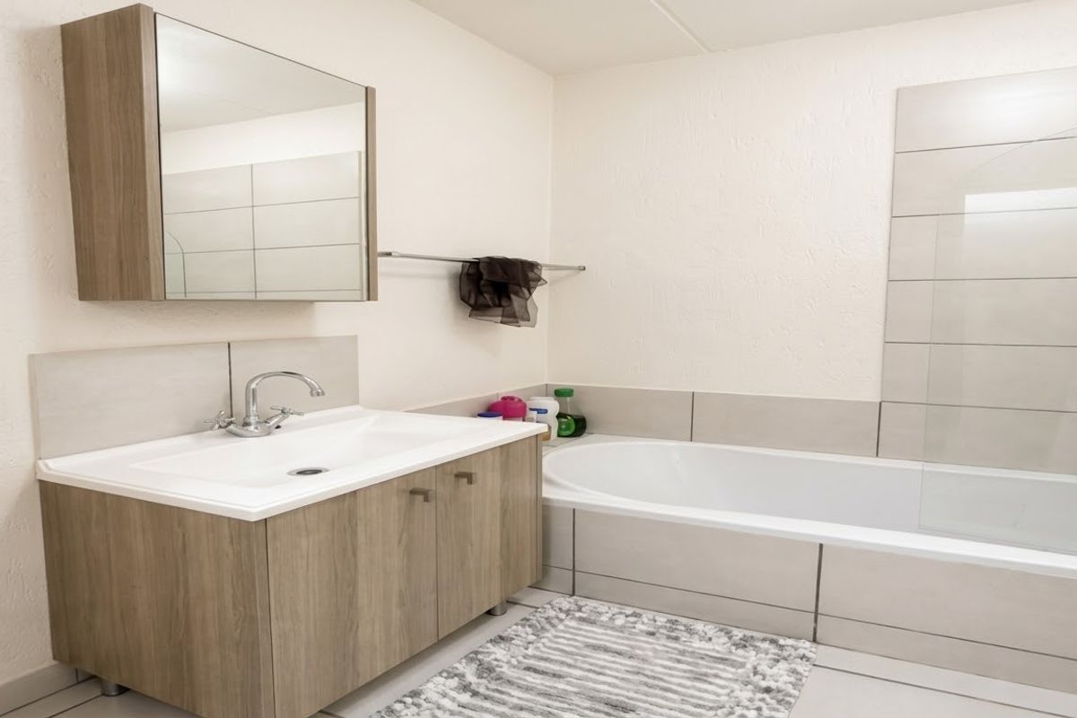 A clean, modern bathroom featuring a wood-finish vanity cabinet, integrated basin, and a bathtub with a glass shower screen and tiled surround.