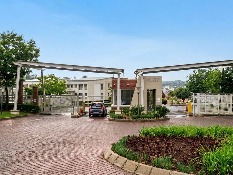 The secure entrance to Alathea Village, featuring a gated access point, guard house, and well-maintained paved driveway with landscaped surroundings.