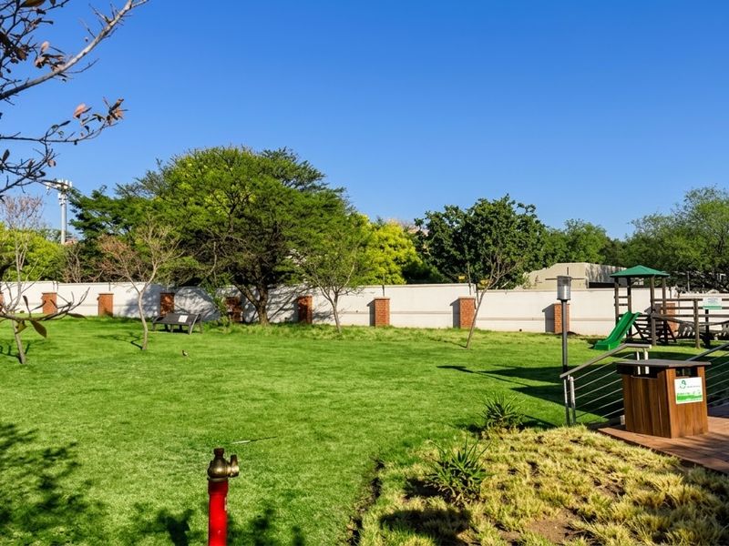 A well-maintained communal garden area featuring a manicured lawn, perimeter wall, paved patio section, and playground equipment.