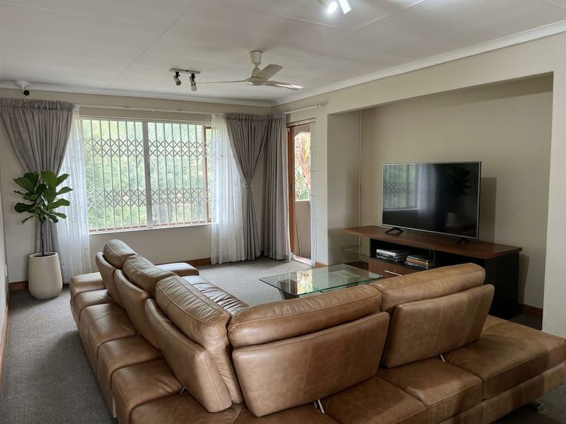 A comfortable living room featuring carpet flooring, a ceiling fan, and large windows fitted with security bars, providing access to an outdoor area.