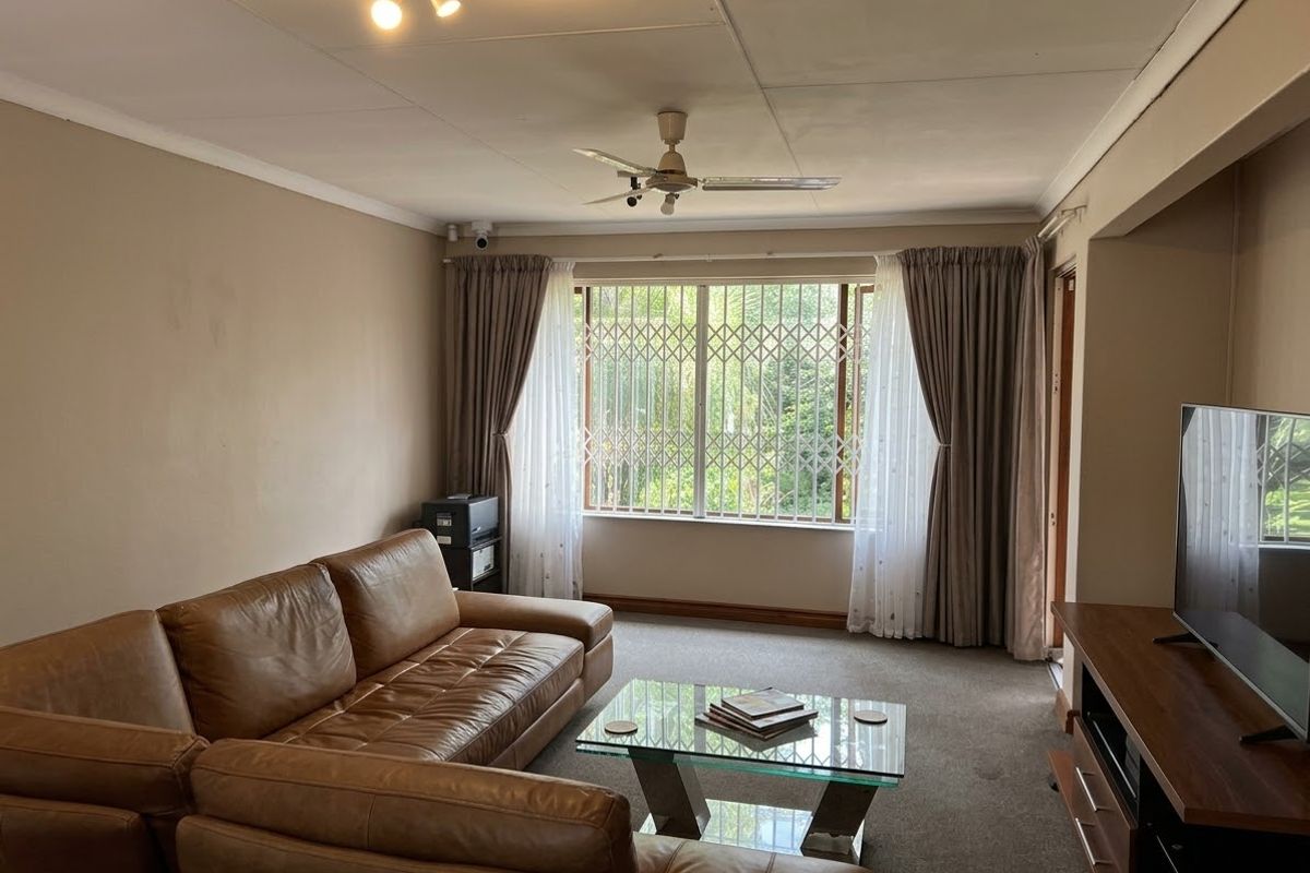 A living room featuring a ceiling fan, carpeted flooring, and a large window fitted with a security gate.