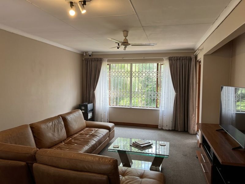 A living room featuring a ceiling fan, carpeted flooring, and a large window fitted with a security gate.