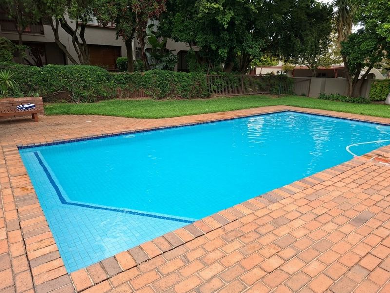 A well-maintained swimming pool area featuring brick paving, a manicured lawn, and secure perimeter fencing within a landscaped garden setting.