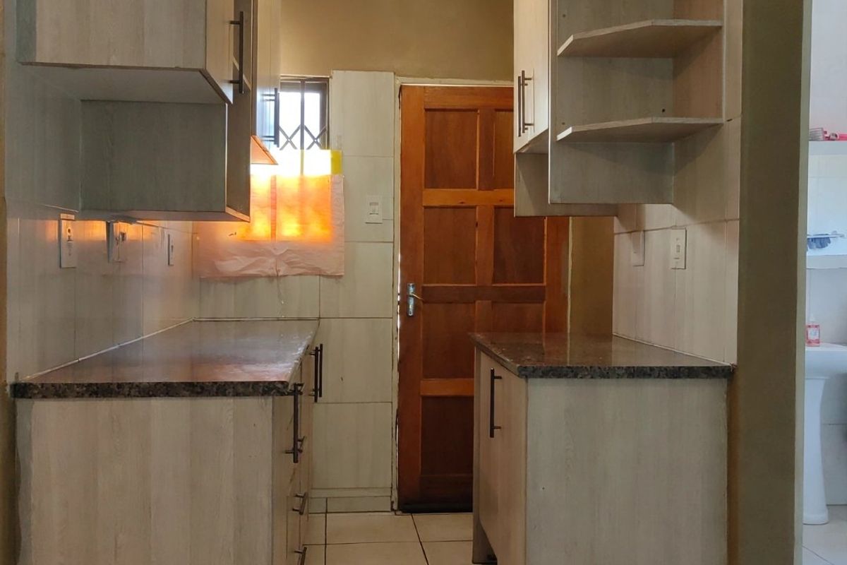A functional galley-style kitchen featuring light wood-finish cabinetry, granite countertops, tiled flooring, and a direct access door to the exterior.