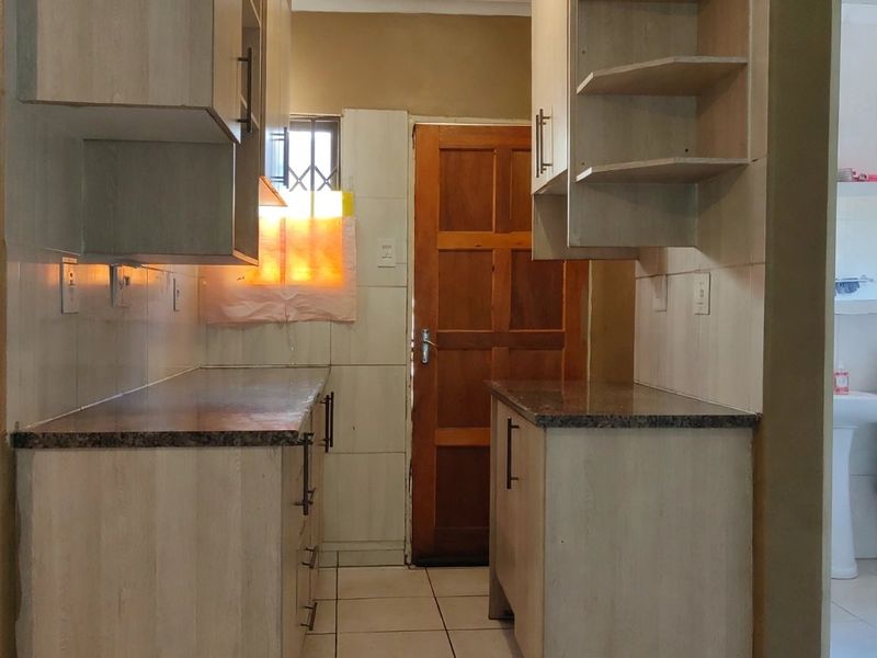 A functional galley-style kitchen featuring light wood-finish cabinetry, granite countertops, tiled flooring, and a direct access door to the exterior.