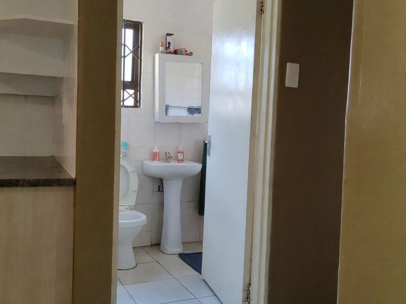 A functional bathroom featuring a pedestal basin, toilet, and tiled flooring, accessed via a hallway with built-in storage shelving.