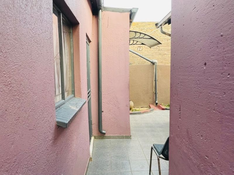 A narrow rear exterior walkway featuring tiled flooring, a downpipe, and a window with security bars, leading to a small paved courtyard area.