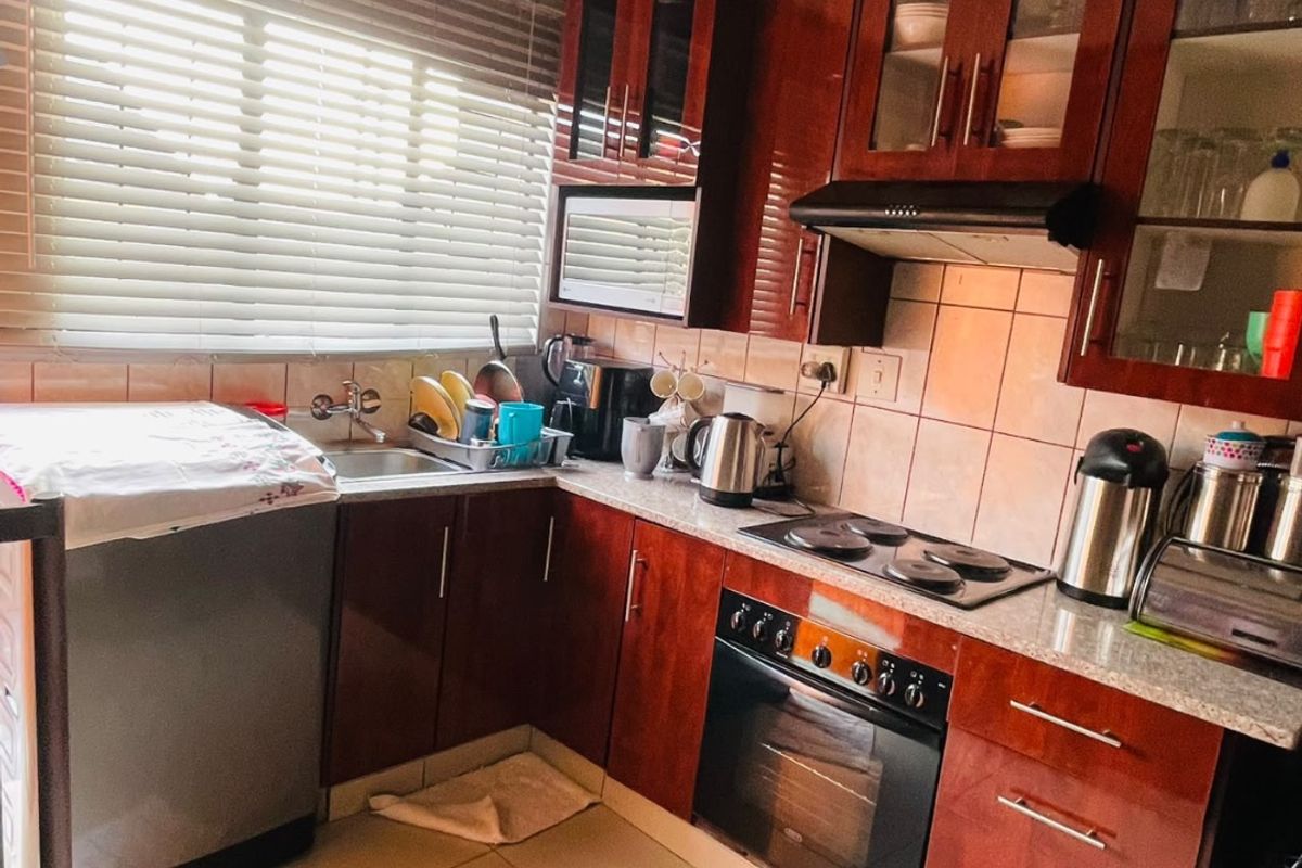 A functional kitchen featuring dark wood cabinetry, tiled backsplash, built-in oven with hob, and a stainless steel sink under a window with blinds.