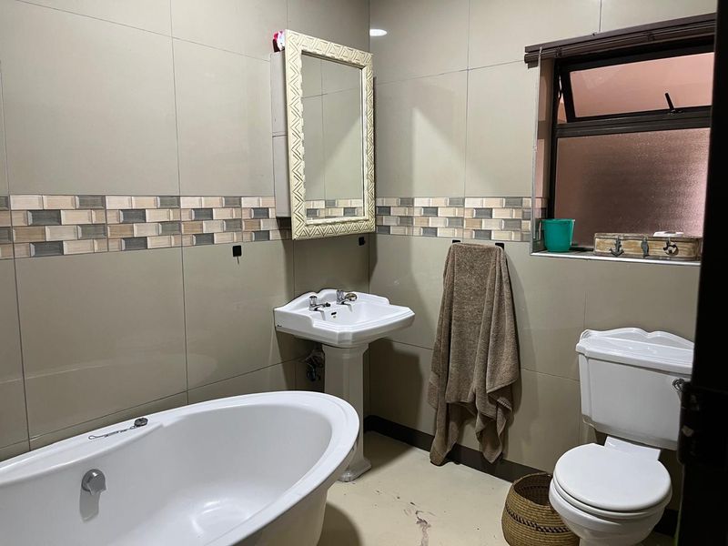 A functional bathroom featuring a pedestal basin, freestanding bathtub, and toilet with neutral wall tiling and a window for natural light.