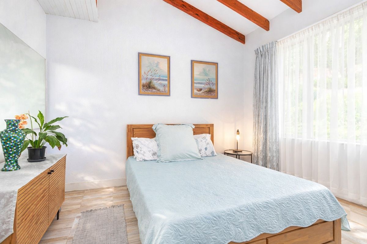 A bright bedroom featuring exposed timber ceiling beams, tiled flooring, and a large window allowing for ample natural light.