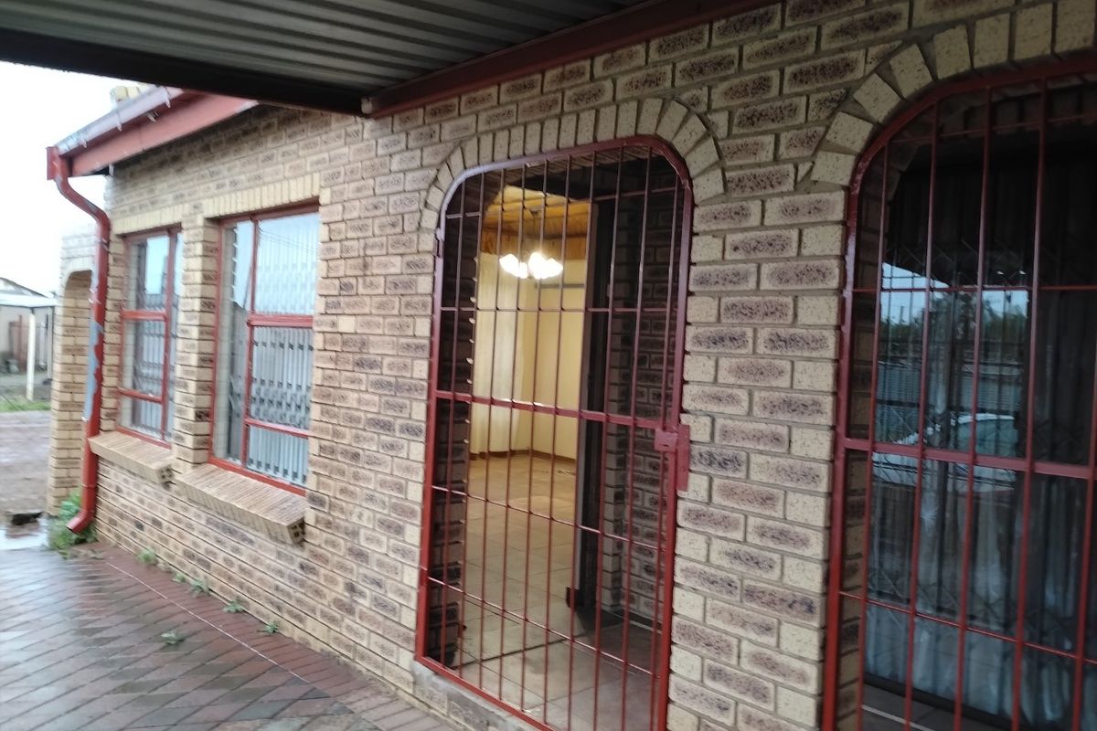 Rear exterior view of a face brick house featuring a paved patio area, metal roof covering, and security gates over the entrance and windows.