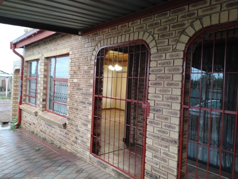 Rear exterior view of a face brick house featuring a paved patio area, metal roof covering, and security gates over the entrance and windows.