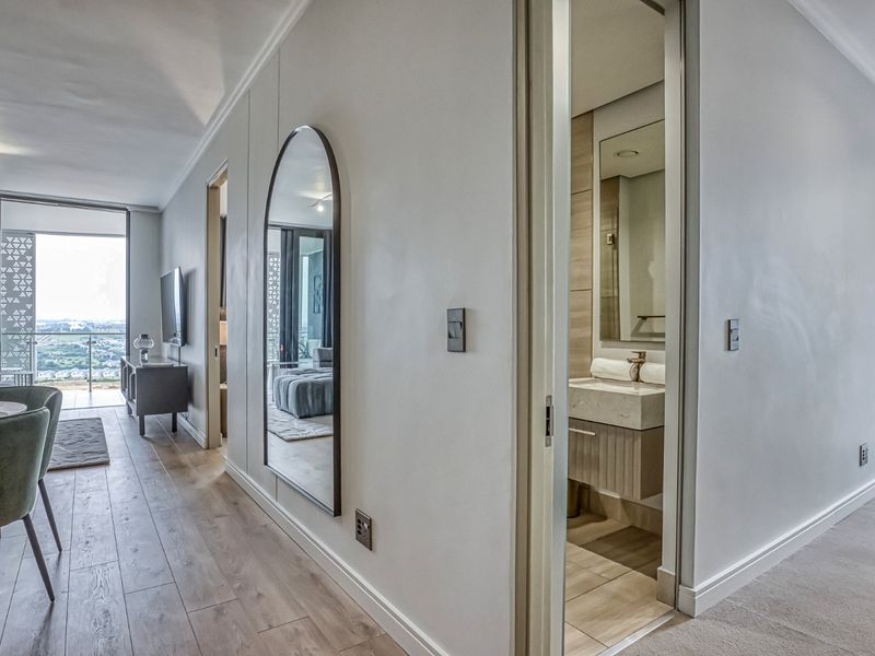 A modern hallway featuring laminate flooring, recessed lighting, and a direct view into a well-appointed bathroom with a stone vanity.