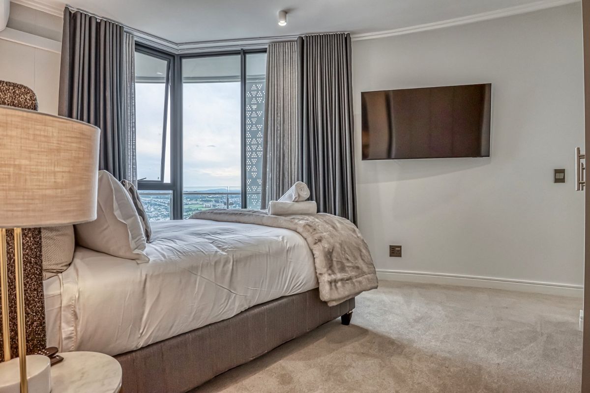A modern, well-maintained bedroom featuring floor-to-ceiling windows with expansive views, neutral carpeting, and a wall-mounted television.