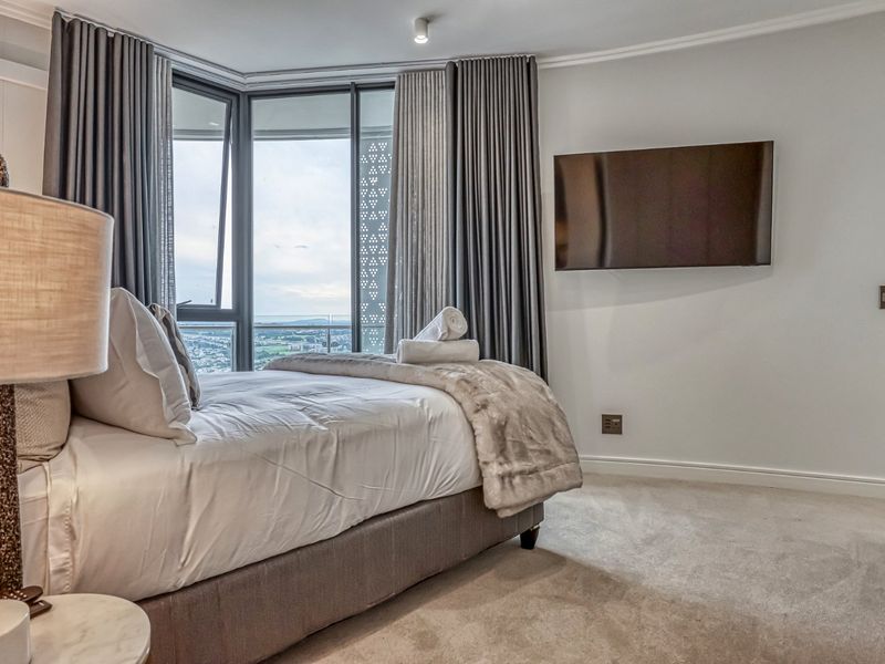 A modern, well-maintained bedroom featuring floor-to-ceiling windows with expansive views, neutral carpeting, and a wall-mounted television.