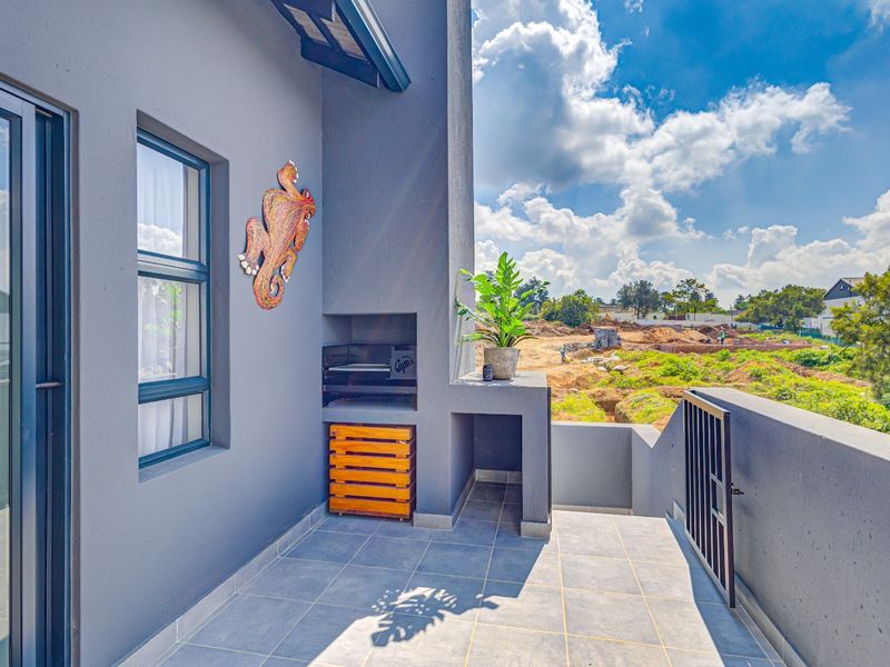 A modern covered balcony featuring a built-in braai, tiled flooring, and a metal security gate, overlooking an adjacent development site.