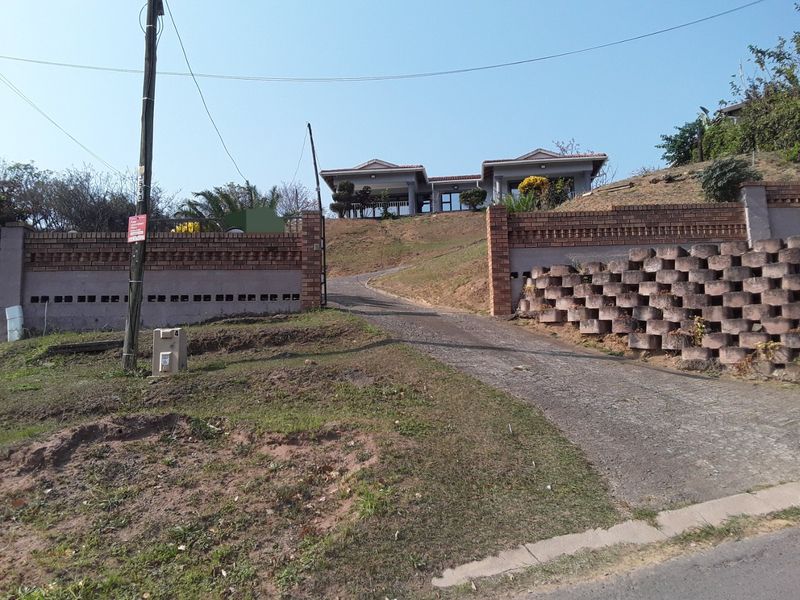Front view of the property featuring a long concrete driveway, brick boundary walls, and a raised house structure with a tiled roof.