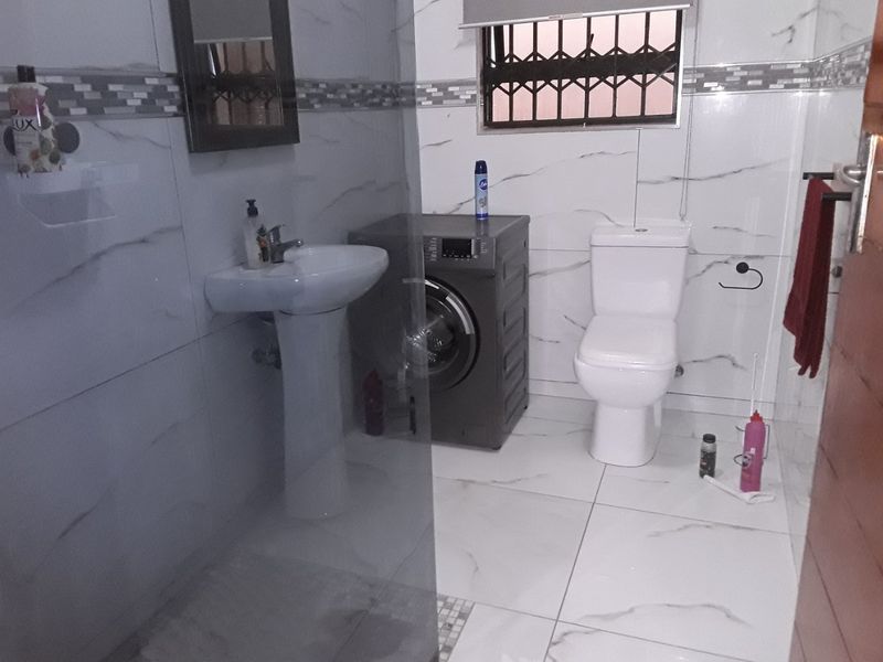 A functional bathroom featuring a pedestal washbasin, toilet, glass shower screen, and integrated laundry space with tiled finishes throughout.