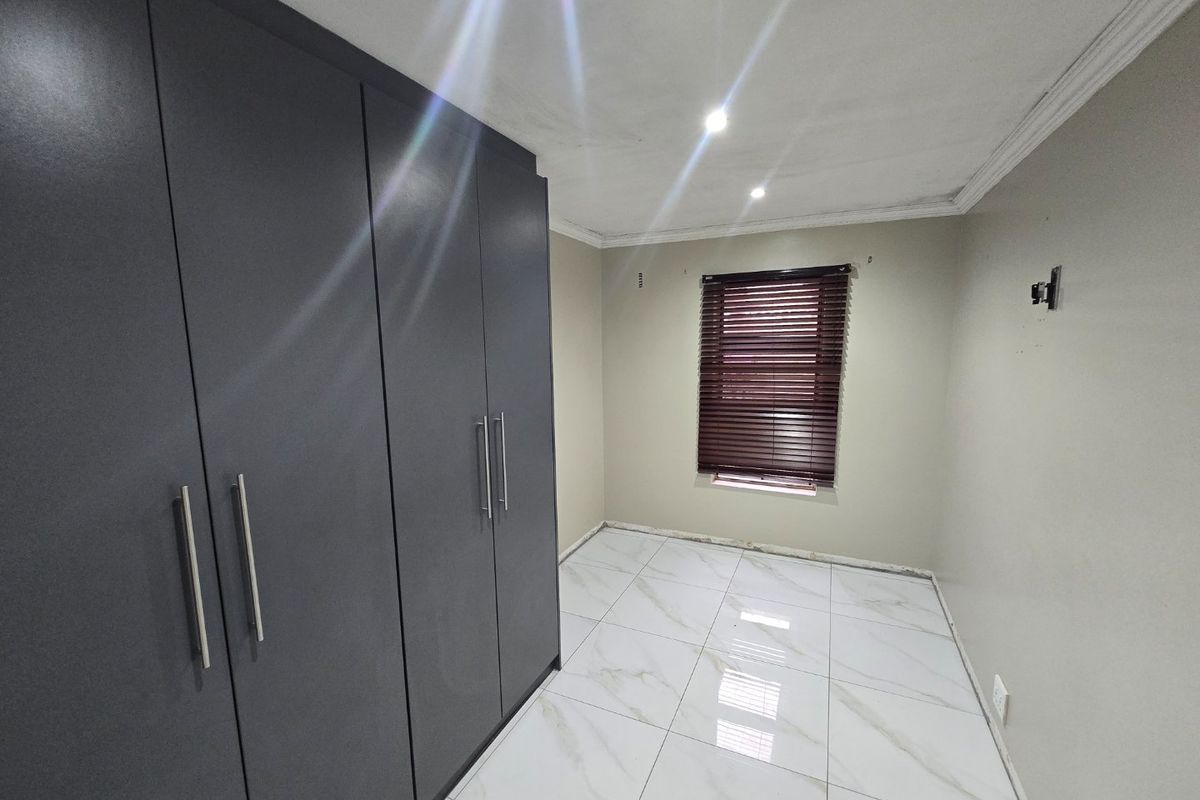 An empty bedroom featuring modern dark grey built-in cupboards, white marble-look tiled flooring, and venetian blinds. The room requires finishing touches like skirting boards.