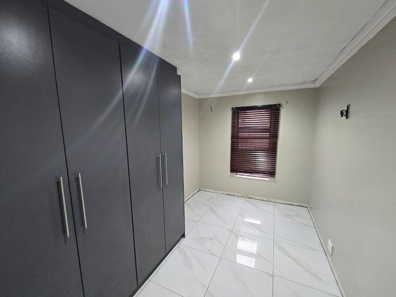 An empty bedroom featuring modern dark grey built-in cupboards, white marble-look tiled flooring, and venetian blinds. The room requires finishing touches like skirting boards.