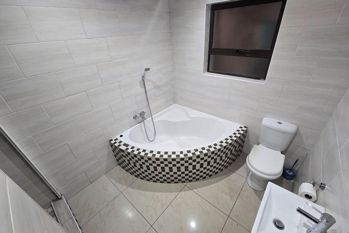A clean, modern bathroom featuring a corner bathtub with decorative checkered tiling, a white toilet, and a wall-mounted basin with neutral wall tiles.
