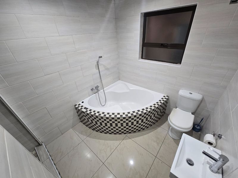 A clean, modern bathroom featuring a corner bathtub with decorative checkered tiling, a white toilet, and a wall-mounted basin with neutral wall tiles.