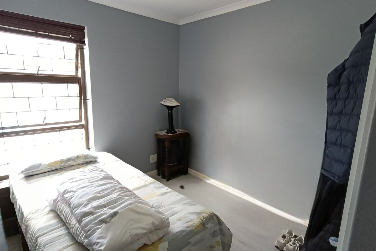 A functional bedroom featuring laminate flooring, neutral grey walls, and a window fitted with security bars.