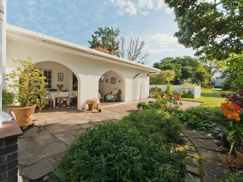 A charming covered patio featuring arched masonry openings, white painted brickwork, and stone paving, overlooking a well-maintained garden with a picket fence.