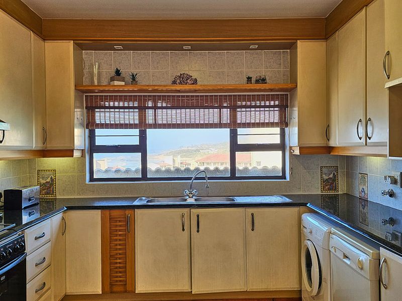 A functional kitchen featuring built-in cabinetry, dark countertops, a stainless steel sink, and integrated laundry plumbing, with a window offering a coastal view.