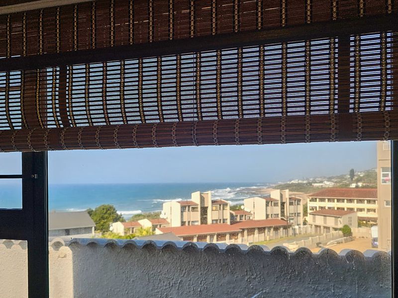 A scenic coastal view overlooking residential buildings and the ocean, framed by a window with a bamboo blind.