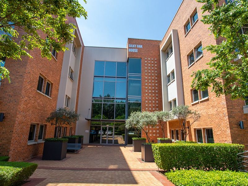 Front entrance view of Silky Oak House, featuring a modern brick and glass facade with a paved courtyard and landscaped garden area.