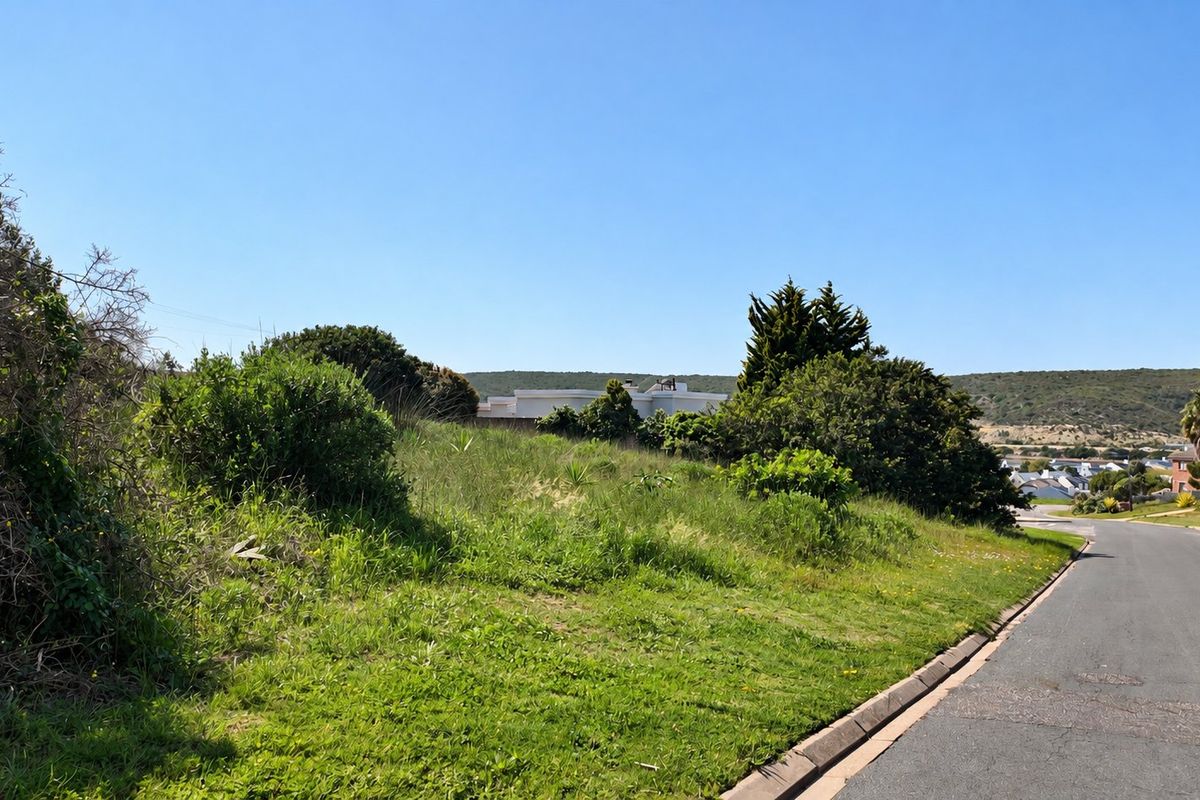 A street-level view of a sloped, undeveloped residential plot featuring natural grass and shrubbery, situated along a paved suburban road.