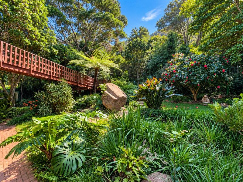 A lush, well-maintained garden featuring a wooden footbridge, brick pathways, and mature landscaping with diverse tropical plants and trees.