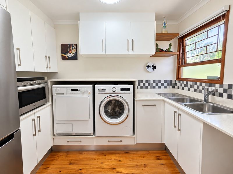 A well-appointed laundry room featuring white cabinetry, timber flooring, a stainless steel double sink, and integrated appliances including an oven and microwave.