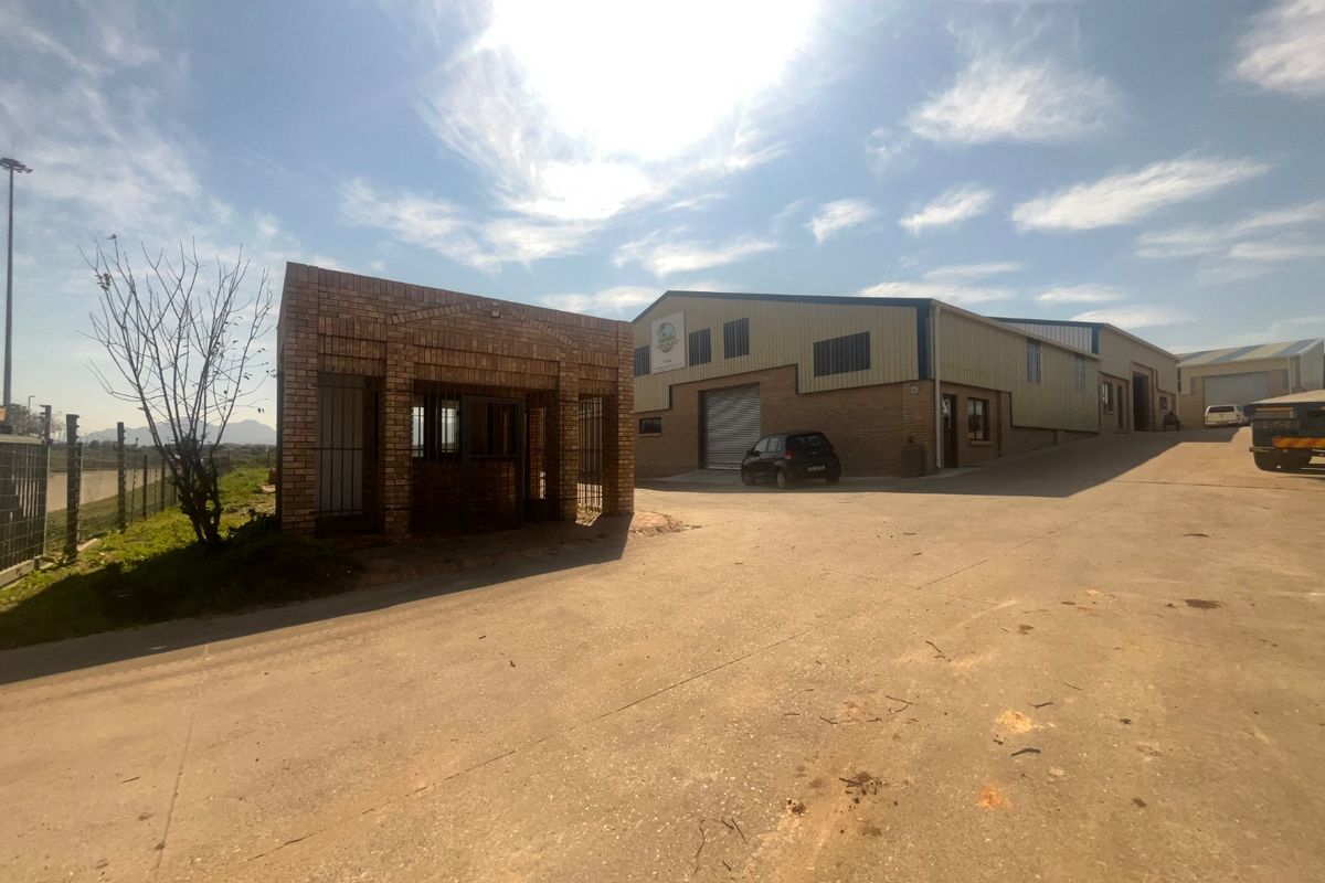 Exterior view of an industrial property featuring a brick security gatehouse, concrete driveway, and a large warehouse building with metal roller shutter doors.