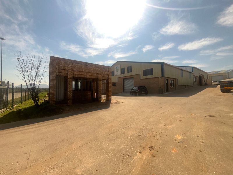 Exterior view of an industrial property featuring a brick security gatehouse, concrete driveway, and a large warehouse building with metal roller shutter doors.