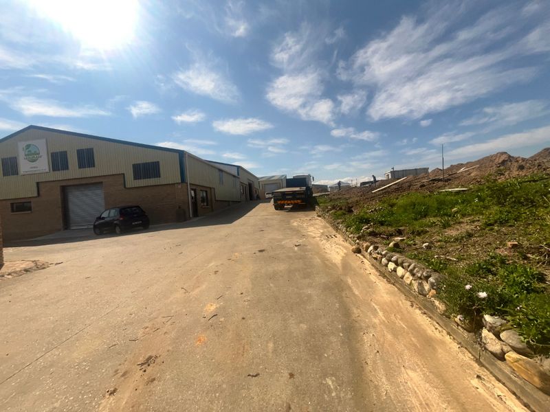Exterior view of an industrial warehouse facility featuring a concrete driveway, roller shutter access, and adjacent earthworks.
