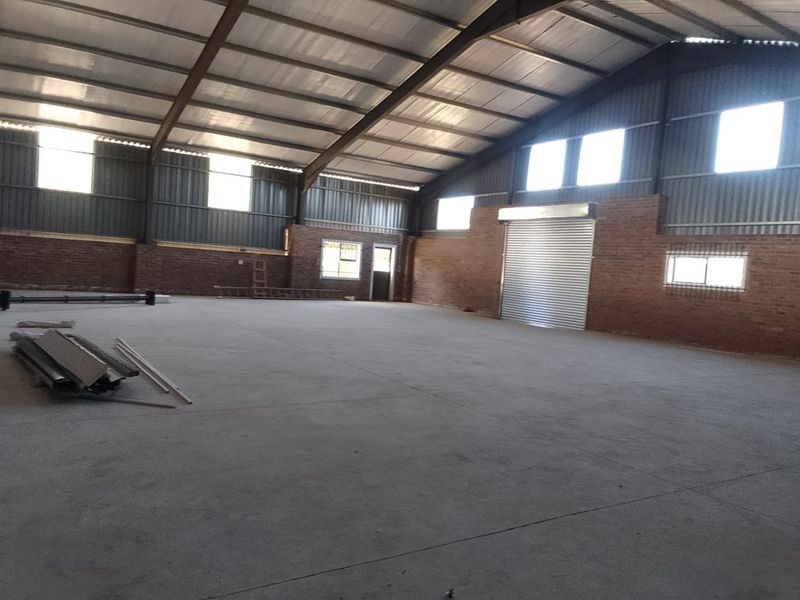 Spacious industrial warehouse featuring concrete flooring, exposed brick walls, and a large metal roller shutter door, suitable for various commercial or storage applications.